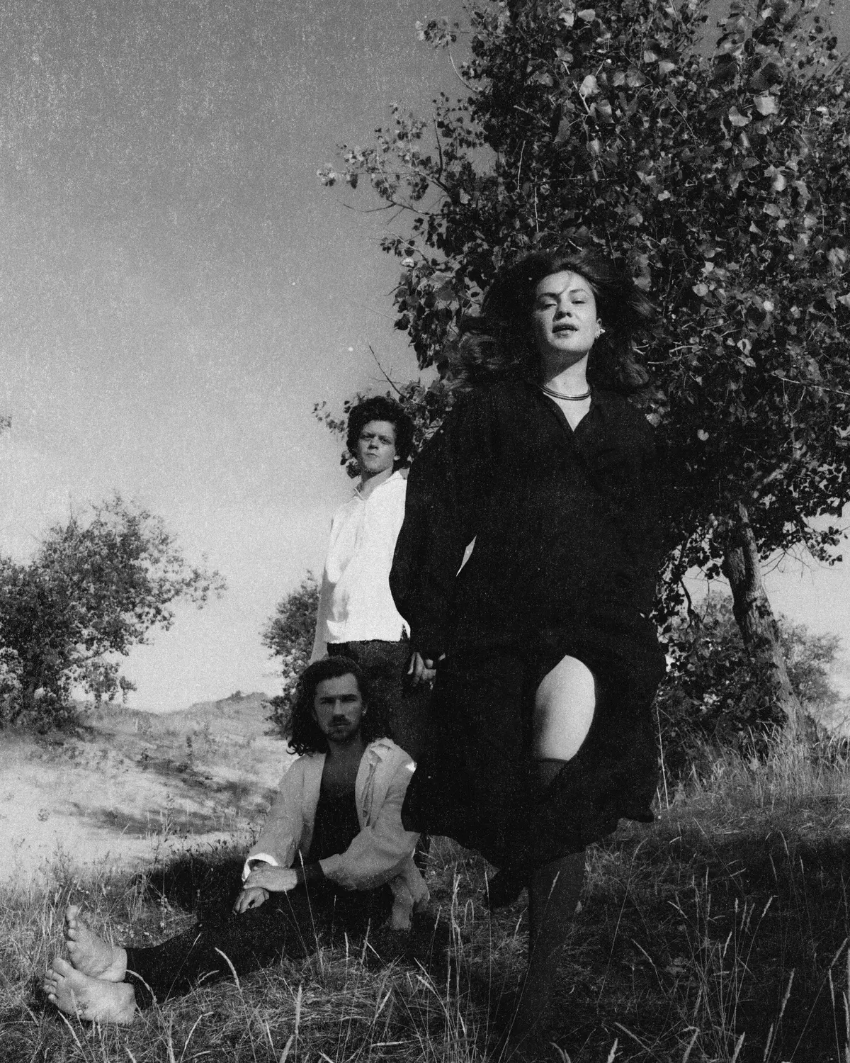 Individual wearing black dress stands in tall grass beside a leafy tree; others visible in the background in a rural landscape.