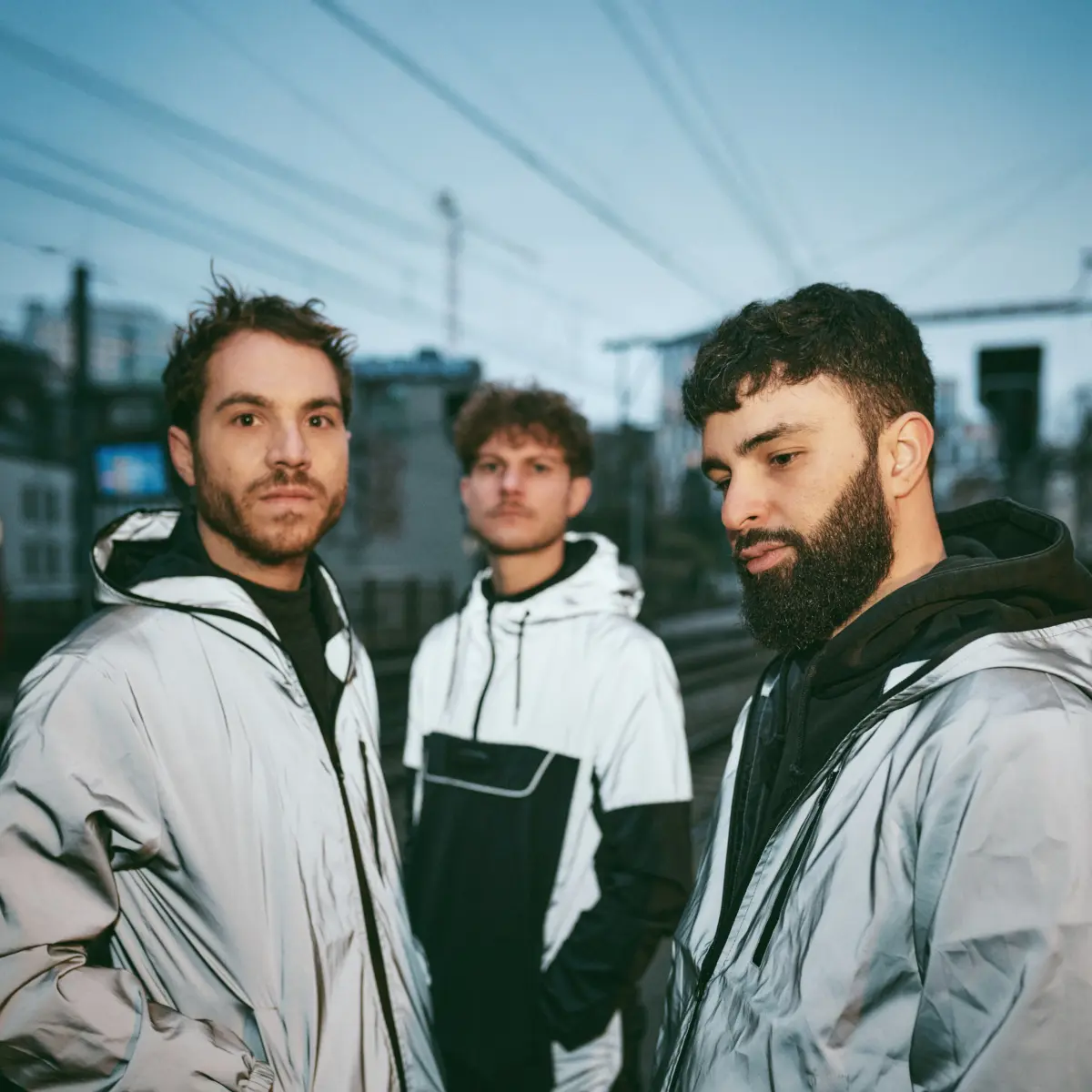 Three young men pose for a selfie in white jackets on an urban rooftop, with power lines in the background.