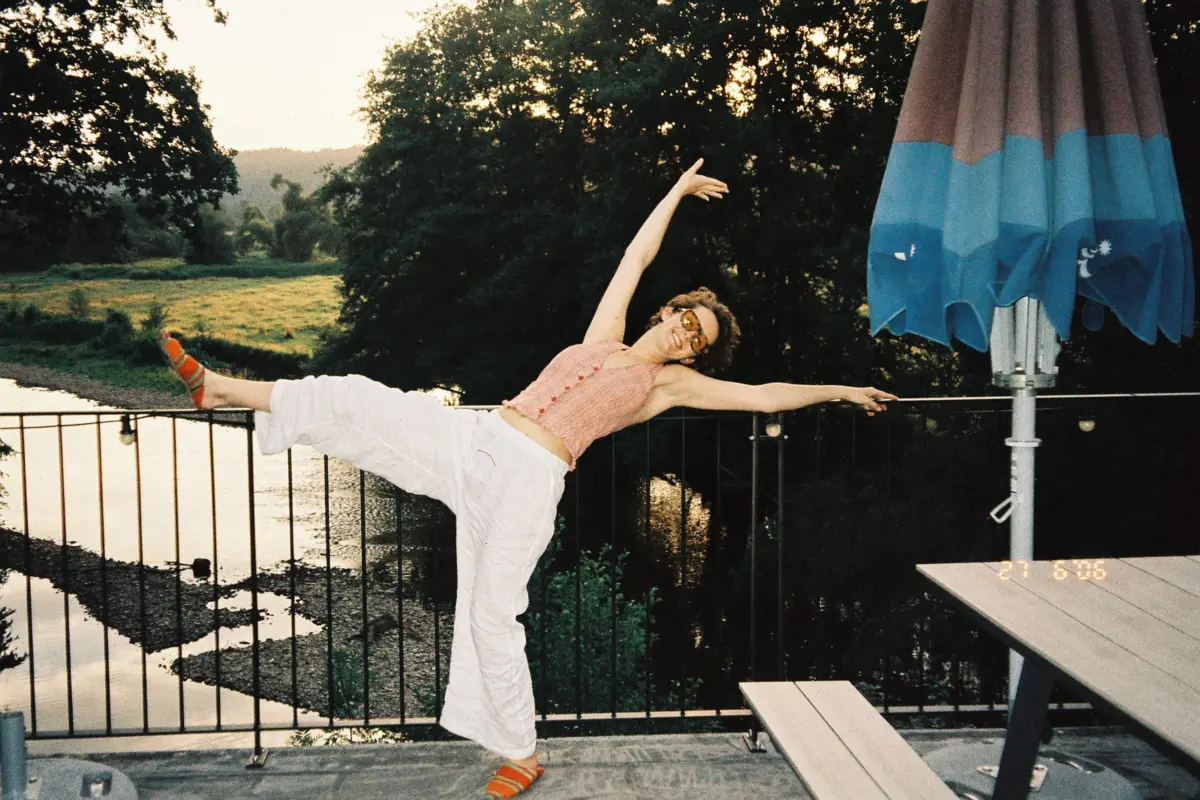 Person in pink sleeveless top and white pants balances on a railing by a lake, with a blue umbrella nearby.