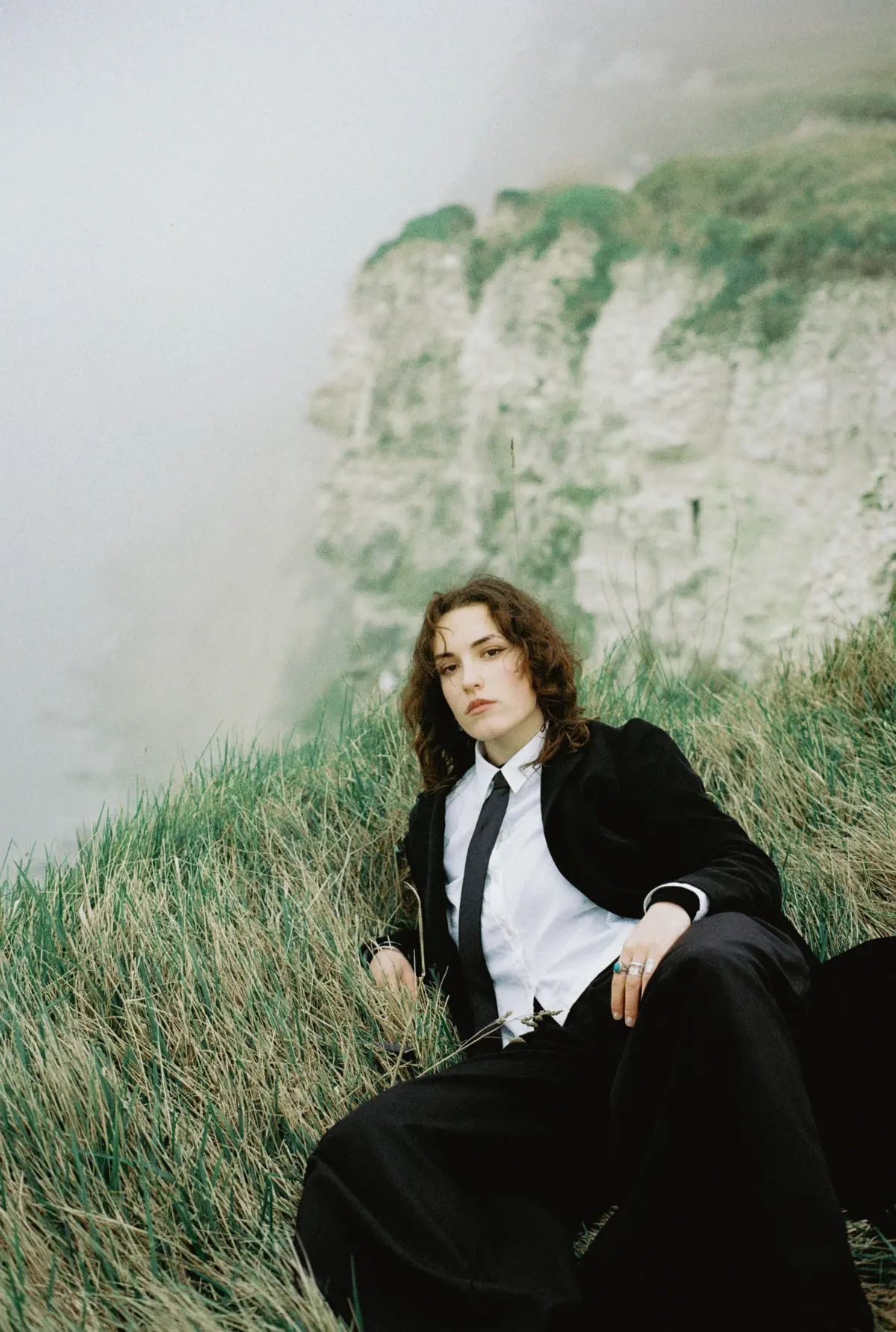 Person with curly hair seated in tall grass on a cliffside, wearing a black suit and white shirt with a black tie; chalky cliffs and sea in background.