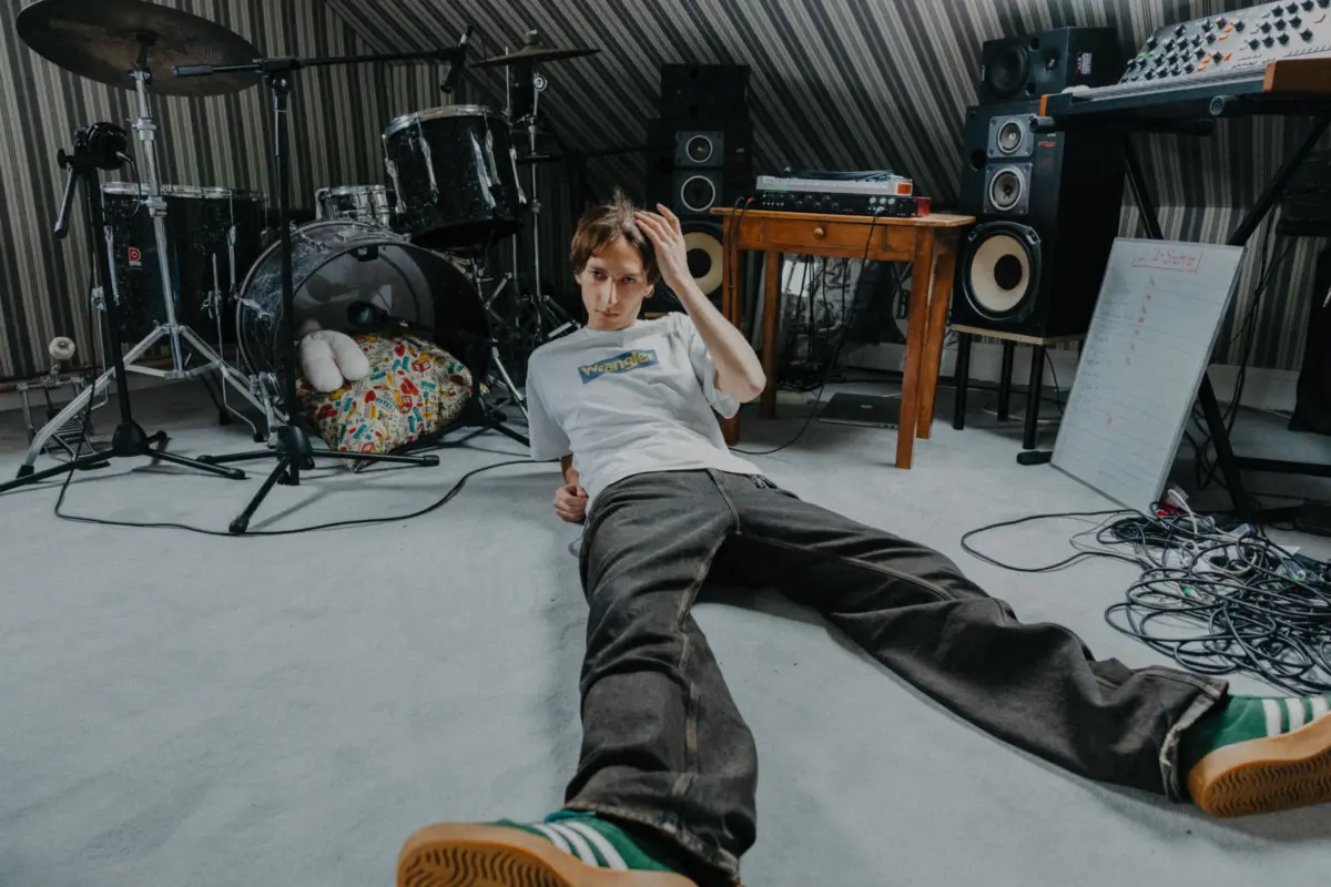 Person lying on the floor of a music studio, hand running through hair, with drums, speakers, keyboard, and cables around.