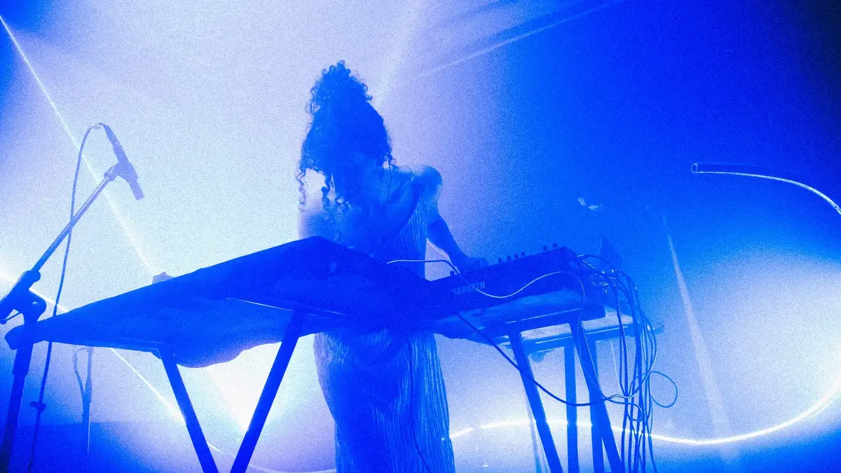 Keyboardist with curly hair performs on a blue-lit stage, surrounded by cables and a microphone stand.