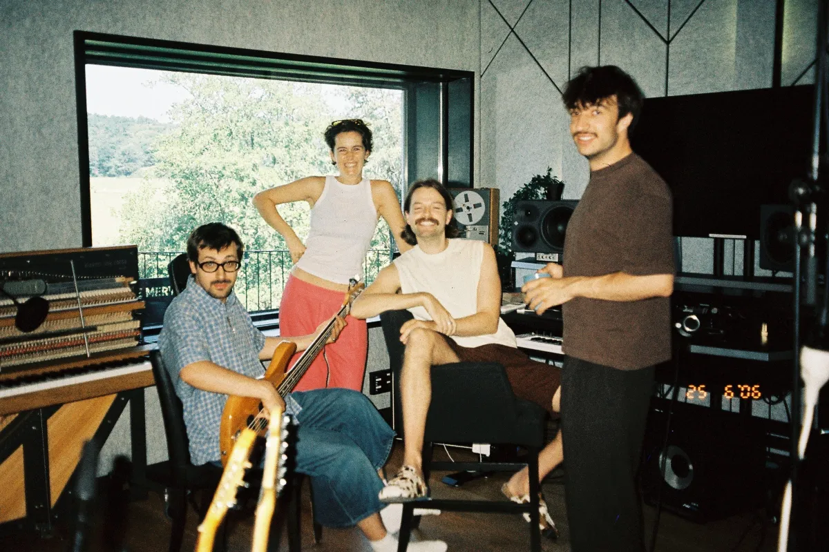 Four men in a recording studio posing; one seated with electric guitar, another at keyboard, others stand by gear.