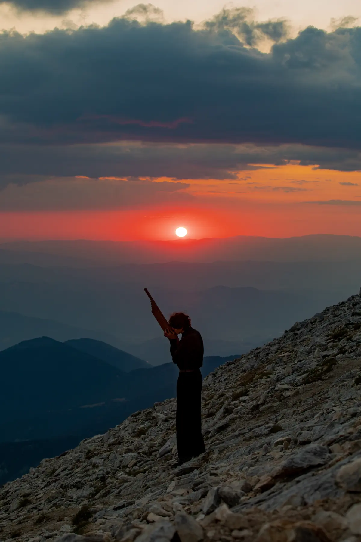 Silhouetted individual on a rocky mountainside at sunset, holding a long, narrow object aloft.