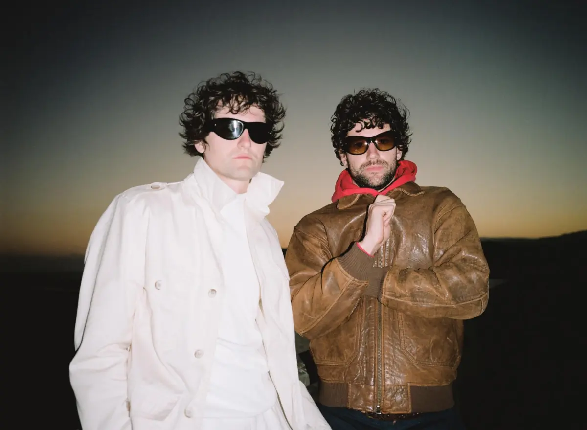 Two men with curly hair and sunglasses pose on a beach at sunset; one wears a white jacket, the other a brown leather jacket with a red hoodie.