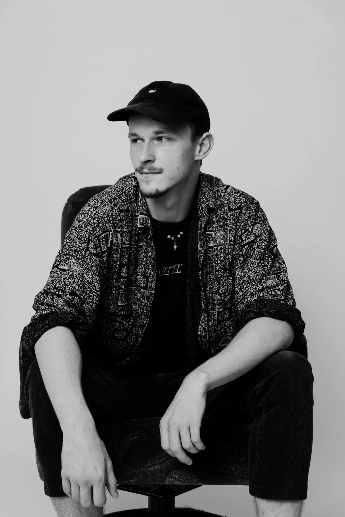 Person wearing a patterned shirt and black cap sits on a stool, looking to the left against a plain wall.