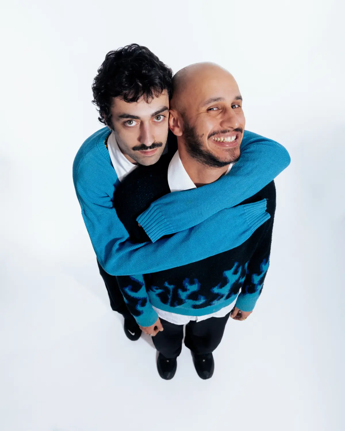Two people hugging from behind in blue sweaters against a white studio backdrop.