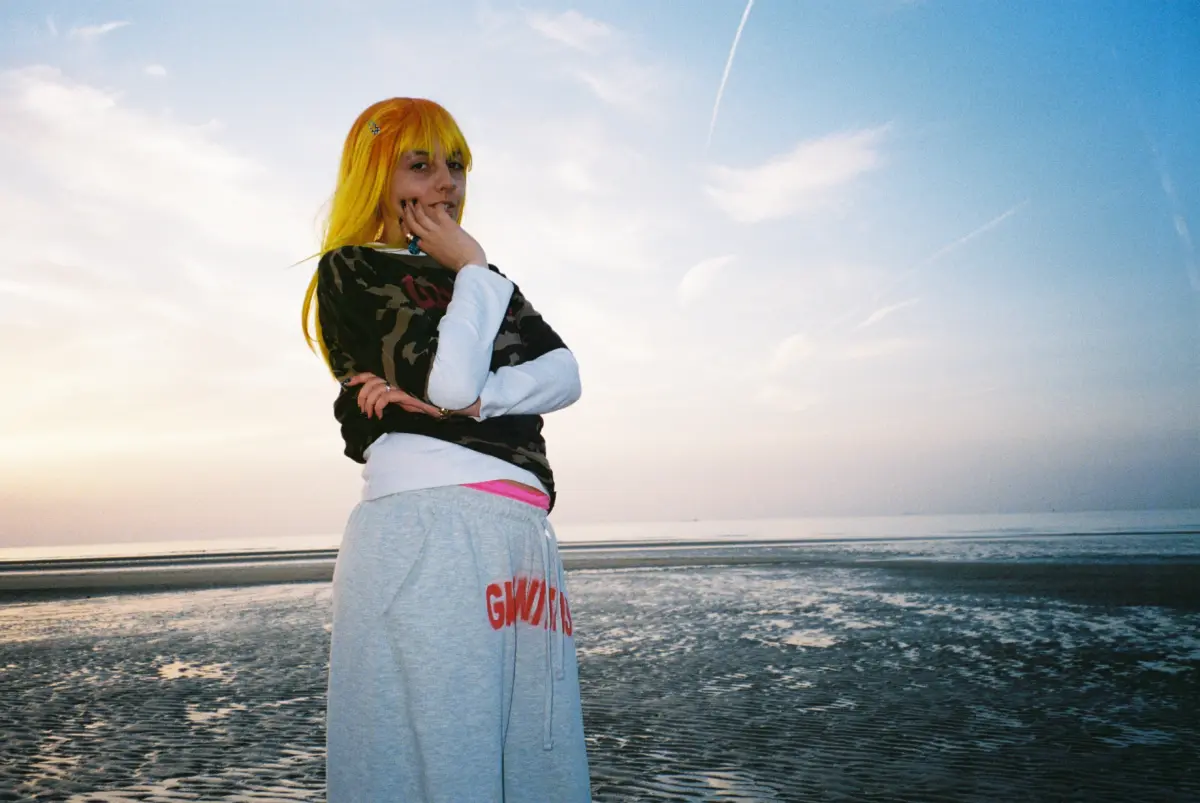 Person with bright yellow hair in camouflage top and gray sweats stands on a beach by the sea at sunset, hand to chin.