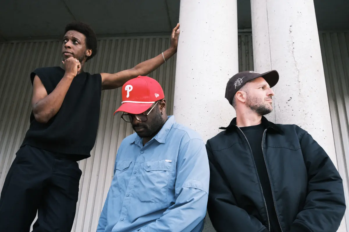 Three men lean against large concrete columns; left man touches chin, others look away.
