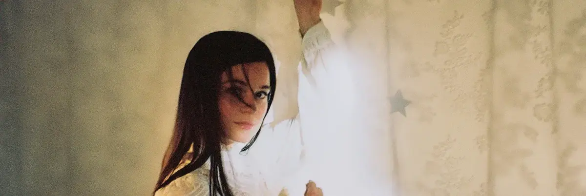 Person with long black hair in a white shirt pulls aside a star-patterned pale curtain in a softly lit room.