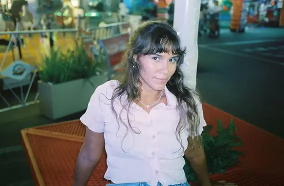 Person with long dark hair in a pink shirt sits leaning against a white post on an orange outdoor bench in a nighttime street market.