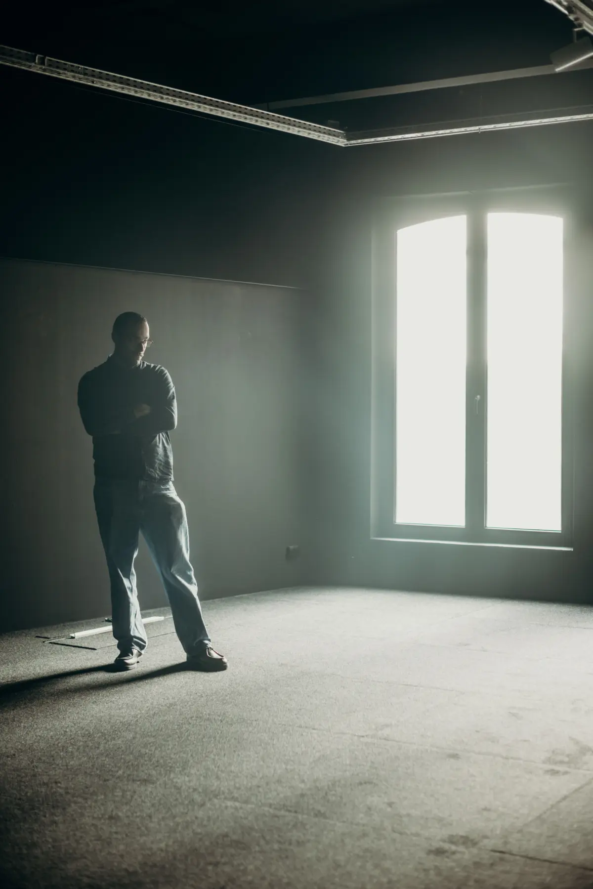 Adult person standing with arms crossed in a dim, empty room with bare walls and a bright window.